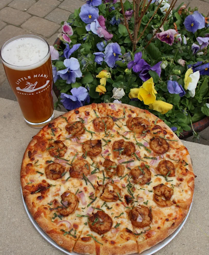 Shrimpalicious pizza and a pint of beer from Little Miami Brewing Company beside a flower pot on an outdoor table.