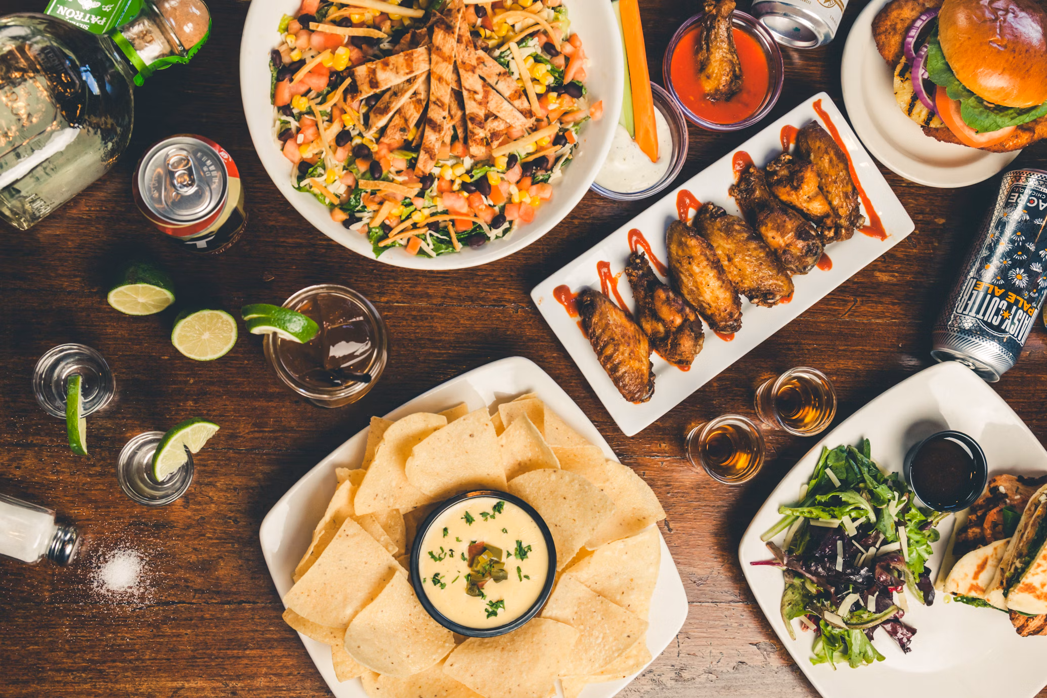 An overhead photo of many dishes- nachos, wings, cans of beer, salad etc