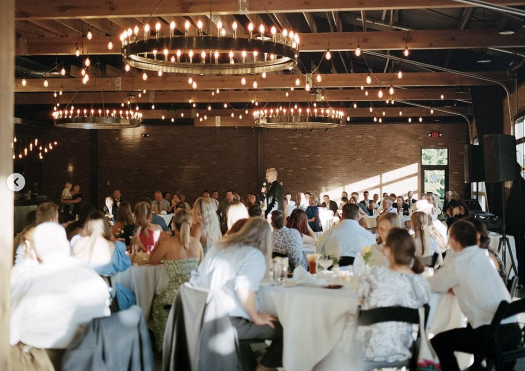 Wedding Reception at Little Miami This image captures a dream wedding reception at Little Miami Brewery in Milford, Cincinnati. The rustic-chic venue features exposed wooden beams, brick walls, and is beautifully lit by circular chandeliers and strings of Edison bulbs. Guests are seated at round tables with white tablecloths, focused on a central figure giving a speech.