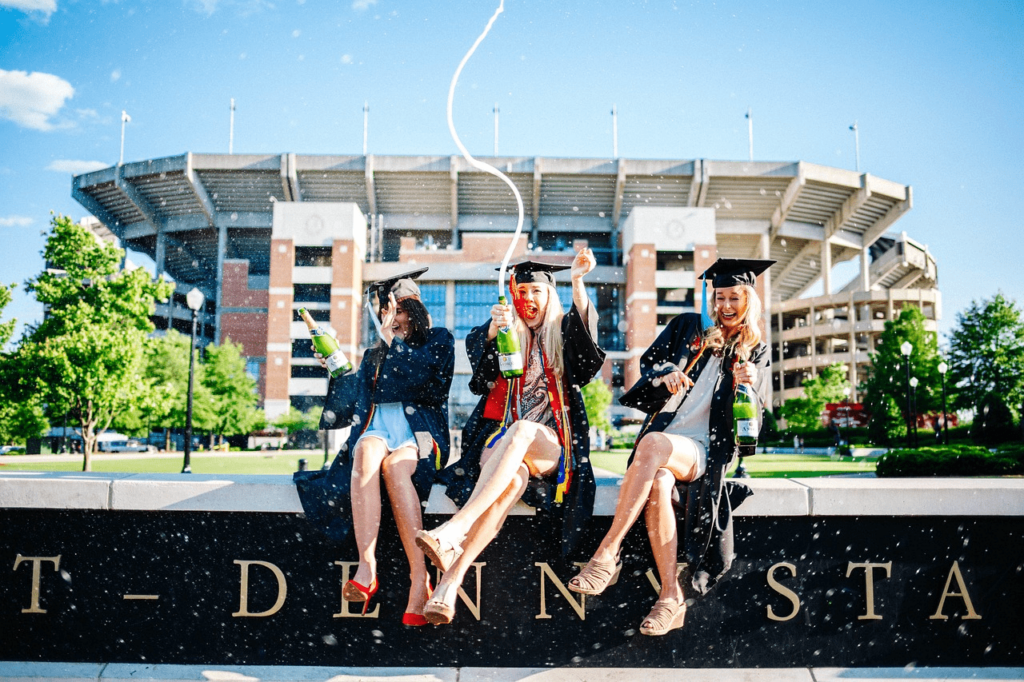 three excited graduates sitting on a low wall The image shows three excited graduates sitting on a low wall in front of a stadium, dressed in black graduation gowns and caps, joyfully celebrating their achievement. Each is holding a bottle of champagne, with the bottles spraying in the air, capturing the exuberant moment. The sky is clear and sunny, adding to the festive atmosphere.