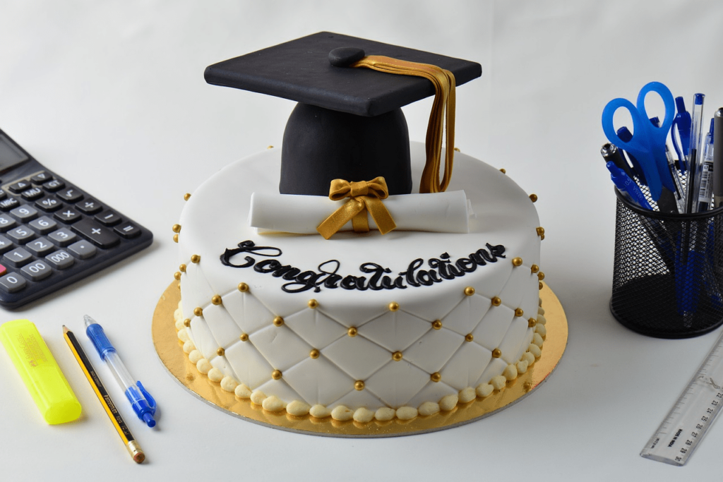 white graduation cake A white graduation cake decorated with a black graduation cap, a diploma with a gold bow, and the words "Congratulations." There is a gold band around the bottom of the cake, and it is decorated with small white dots. To the left of the cake are a calculator, a yellow highlighter, a blue pen, and a pencil.