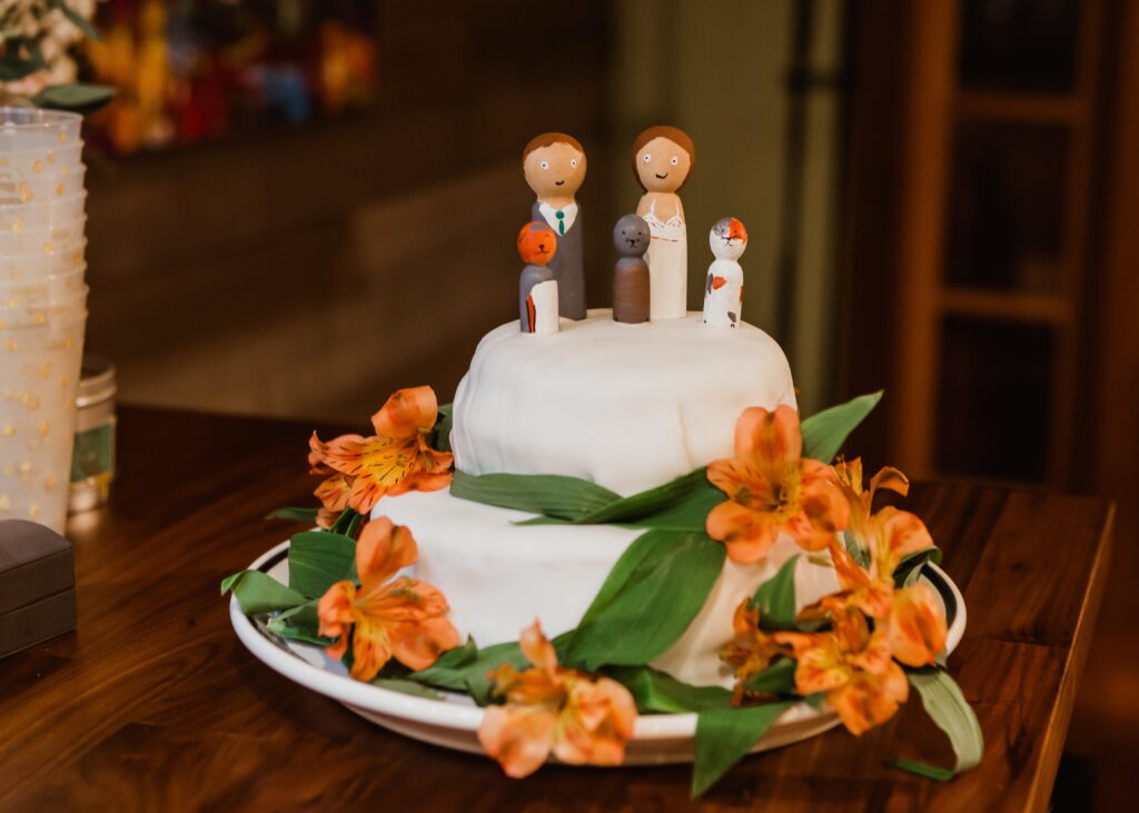 two-tiered wedding cake A two-tiered wedding cake decorated with orange flowers and greenery. The top tier features wooden figurines representing the bride, groom, and their dog.