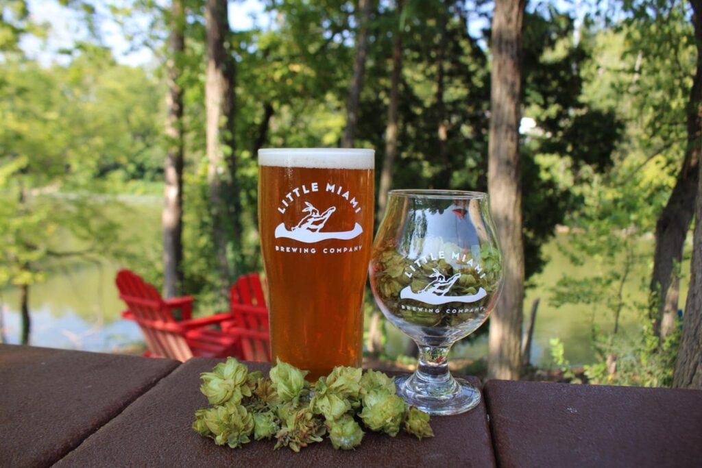 A pint glass of Little Miami Brewing Company beer A pint glass of Little Miami Brewing Company beer with a hop flower and a tulip glass on a table overlooking a lake and trees.