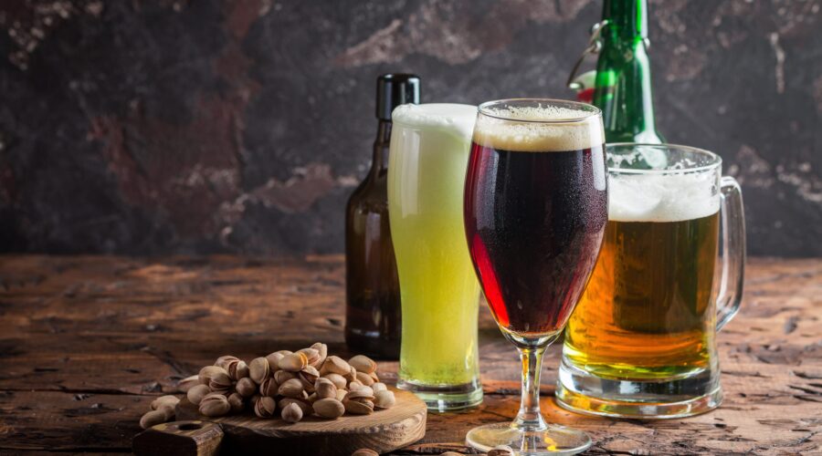 Three different beers with a bowl of pistachios on a wooden table.