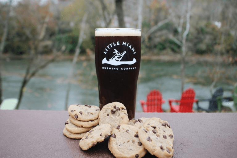 A pint glass filled with dark beer A pint glass filled with dark beer sits on a table next to a pile of chocolate chip cookies, with a scenic river view in the background.