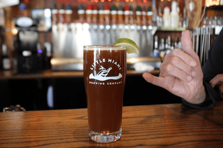 A pint glass filled with dark beer A pint glass filled with dark beer sits on a bar counter with a lime wedge and a hand making the shaka sign in the background.