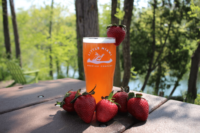 A pint glass filled with golden beer A pint glass filled with golden beer sits on a wooden table outside with a scenic view of the river and several strawberries.