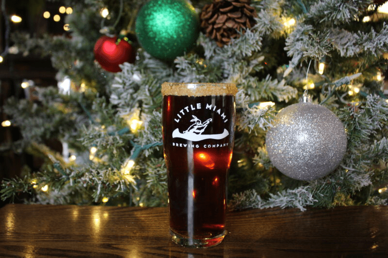 A pint glass A pint glass filled with dark beer sits on a wooden table in front of a decorated Christmas tree.