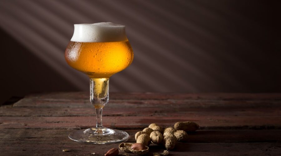 A tulip glass filled with golden Belgian beer, topped with a frothy white head, sits on a rustic wooden table alongside scattered peanuts.