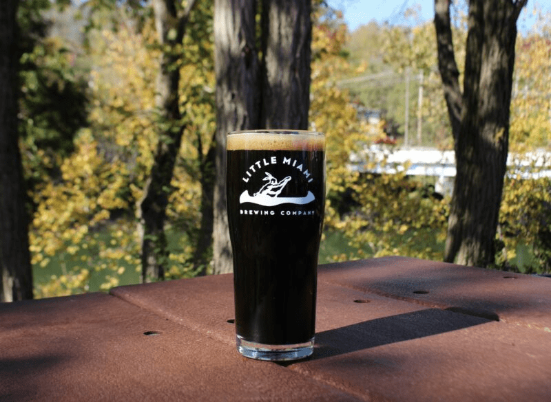 A pint glass filled with dark beer A pint glass filled with dark beer sits on a wooden table outside with a scenic view of the river.