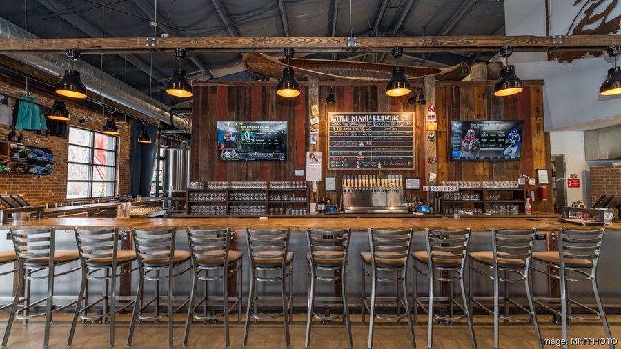 A cozy brewery bar with a rustic wooden design A cozy brewery bar with a rustic wooden design featuring a long wooden countertop lined with high metal chairs. Behind the bar, a chalkboard displays the beer menu from "Little Miami Brewing Co." with taps below it. Two mounted TVs on either side of the chalkboard display sports games. The space is warmly lit with industrial-style pendant lights, and the exposed brick walls and ductwork add to the brewery's inviting and industrial-chic aesthetic.