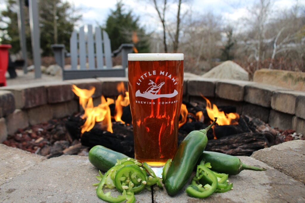 A close-up shot of a pint glass of beer A close-up shot of a pint glass of beer from Little Miami Brewing Company. The glass is placed on a stone ledge in front of a cozy outdoor fireplace. The amber beer has a frothy head, and beside the glass, there are sliced jalapeno peppers, suggesting that the beer flavor is spicy.