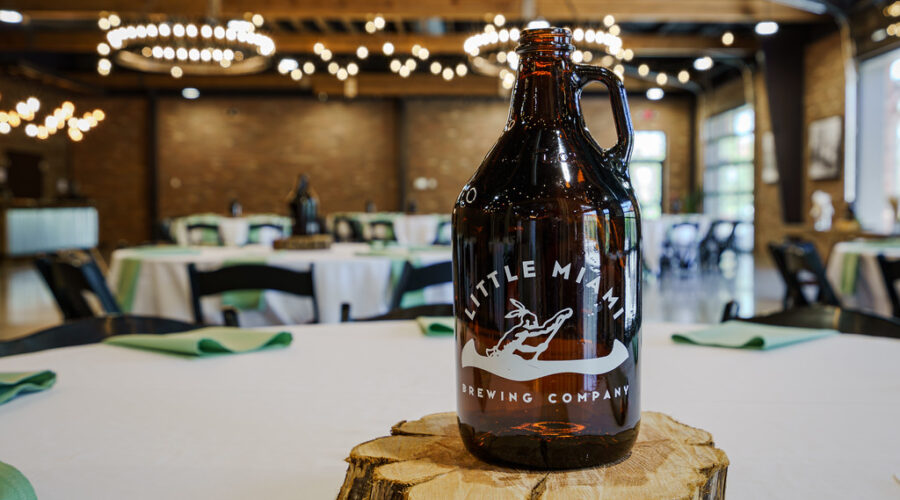 A growler from Little Miami Brewing Company on a wooden slab, with tables and chandeliers in the background.