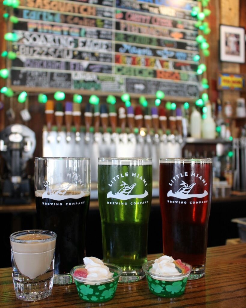 A selection of colorful drinks from Little Miami Brewing Company with green lighting and a chalkboard menu in the background.