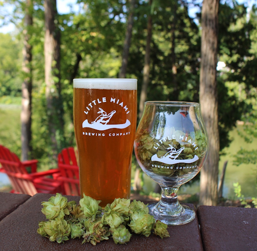 A pint of beer and a glass of beer next to hops, with a scenic view of the Little Miami River in the background.